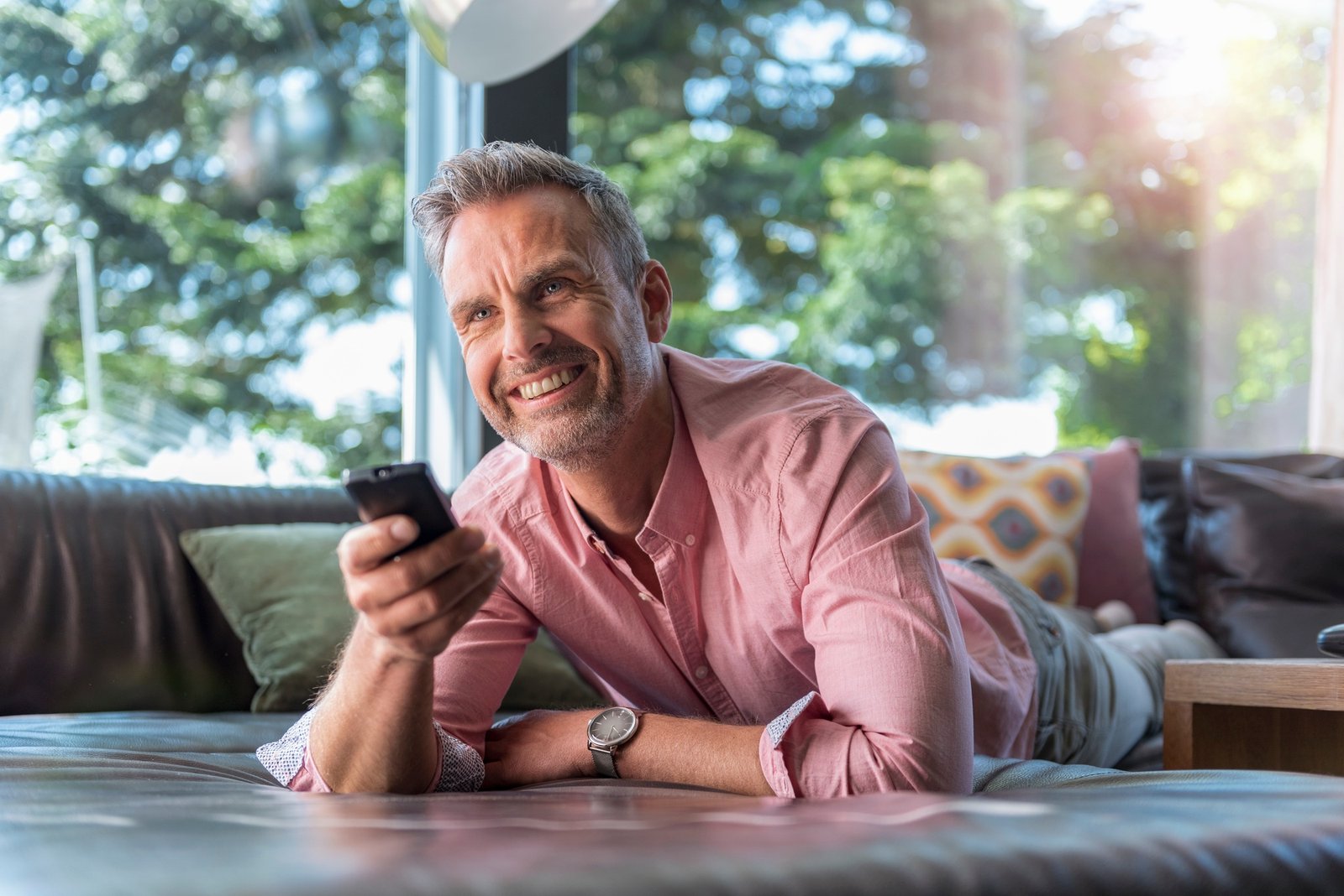 Smiling mature man lying on couch at home using remote control